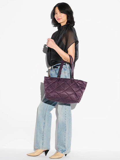 A woman stands sideways, wearing a sheer black blouse, light blue jeans, beige pointed shoes, and carrying the MZ Wallace Medium Madison E-W Tote in Raisin Oxford.
