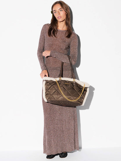A woman in a long-sleeved, floor-length brown dress holds the MZ Wallace Medium Madison E-W Tote in Shearling & Walnut, featuring Italian leather trim, a gold chain handle, and quilted detailing.