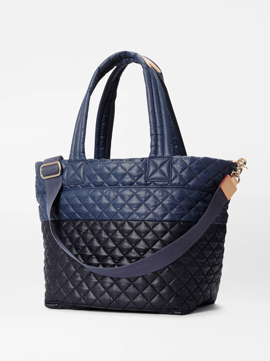 The MZ Wallace Medium Metro Tote Deluxe in Black/Navy Color Block Oxford boasts a sophisticated quilted design, two handles, and an adjustable crossbody strap. The bag features contrasting black and navy sections with gold hardware, providing a blend of style and functionality.