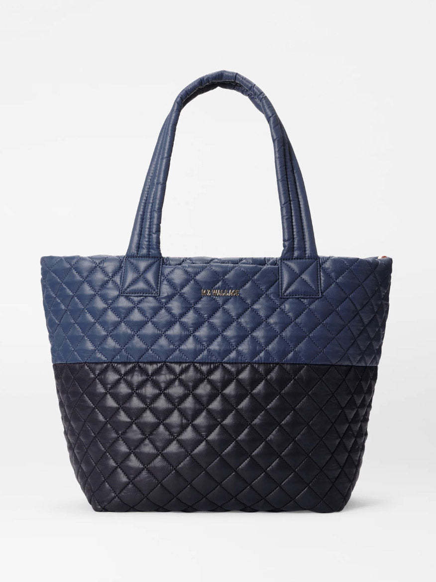 The MZ Wallace Medium Metro Tote Deluxe in Black/Navy Color Block Oxford is a quilted tote bag with two handles, showcasing a striking two-tone design with dark blue on the upper half and black on the lower half against a plain white background. It also includes an adjustable crossbody strap for added convenience.