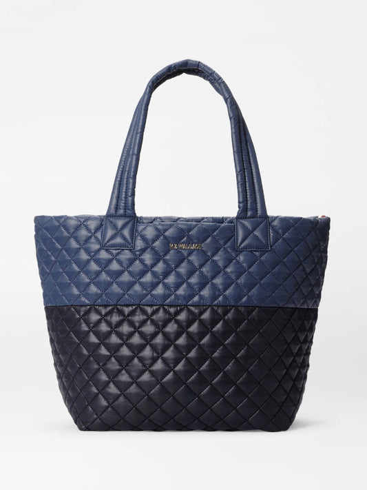 The MZ Wallace Medium Metro Tote Deluxe in Black/Navy Color Block Oxford is a quilted tote bag with two handles, showcasing a striking two-tone design with dark blue on the upper half and black on the lower half against a plain white background. It also includes an adjustable crossbody strap for added convenience.