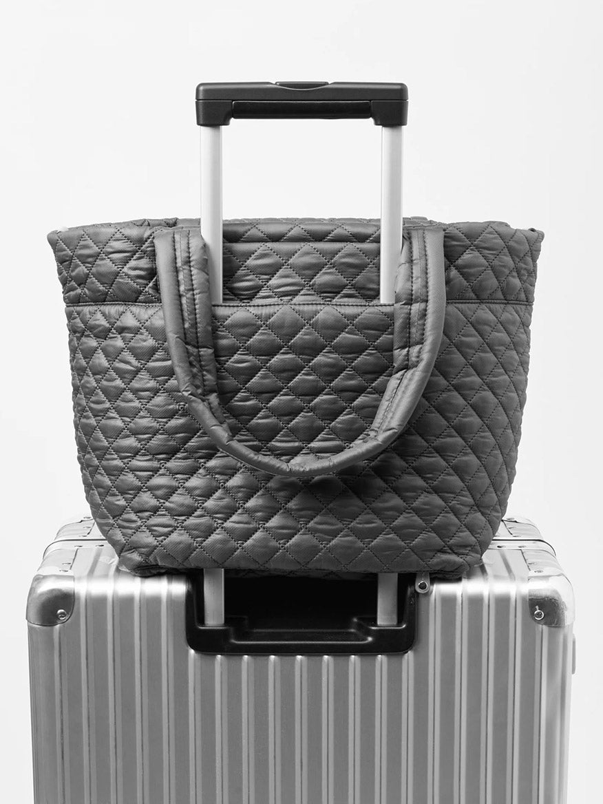 An MZ Wallace Medium Metro Tote Deluxe in Black/Navy Color Block Oxford, complete with an adjustable crossbody strap, is placed on top of a silver hard shell carry-on suitcase with an extended handle.