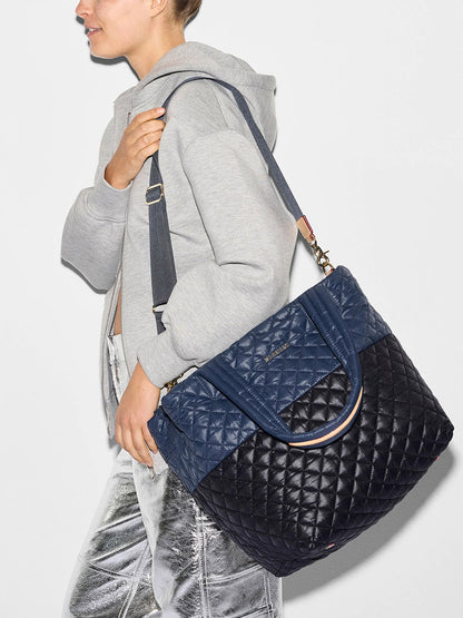 Person wearing a grey hoodie and metallic silver pants, carrying an MZ Wallace Medium Metro Tote Deluxe in Black/Navy Color Block Oxford with an adjustable crossbody strap over their shoulder.