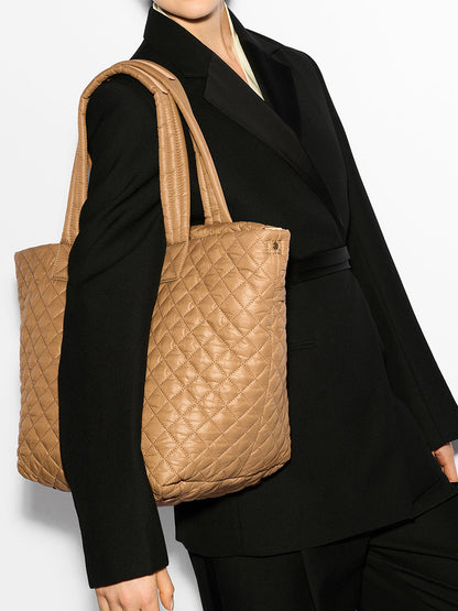 A person in a black blazer holds the stylish MZ Wallace Medium Metro Tote Deluxe in Caramel Oxford with long straps, set against a plain white background.