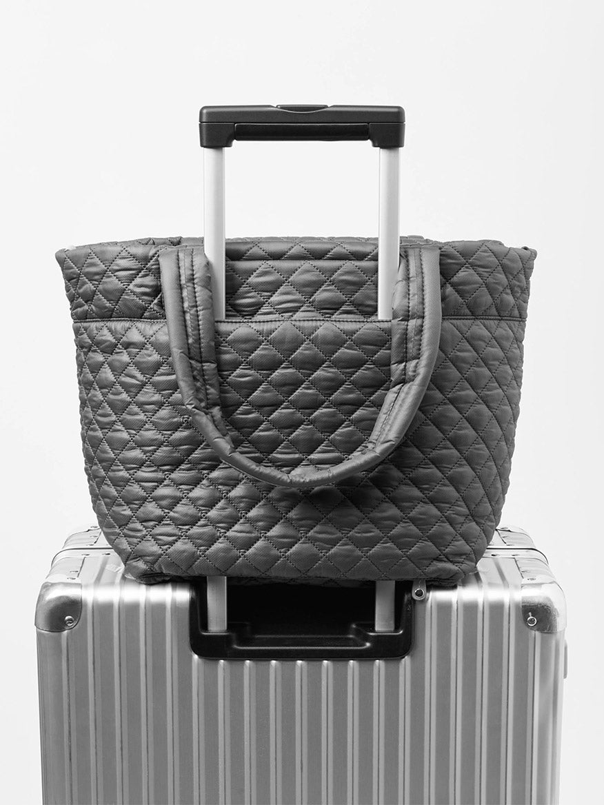 The MZ Wallace Medium Metro Tote Deluxe in Morning Dove Oxford rests atop a hard-shell suitcase, with its luggage sleeve secured over the suitcase handle for convenient travel.