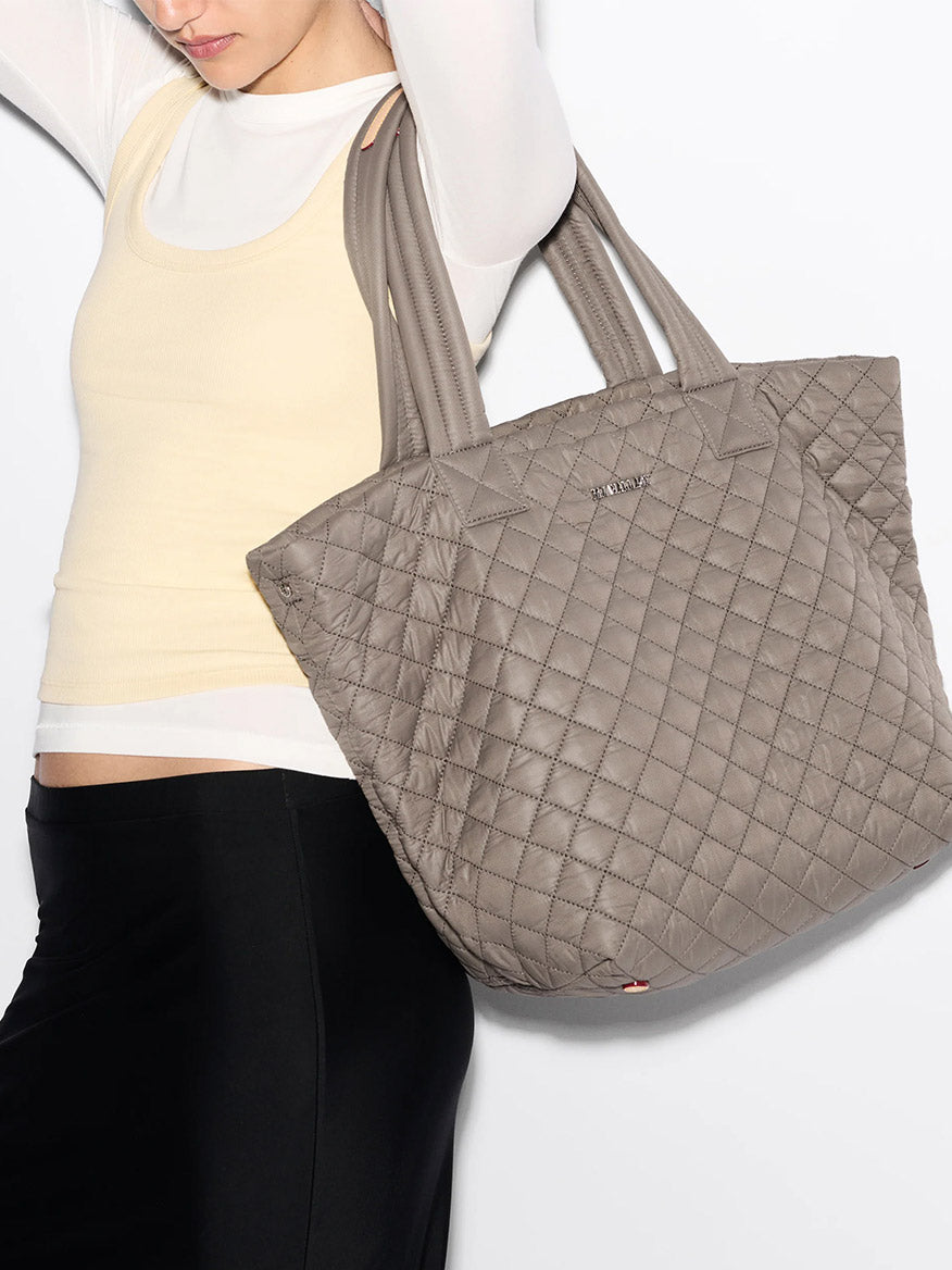 A person wearing a light yellow top and black pants carries the MZ Wallace Medium Metro Tote Deluxe in Morning Dove Oxford with an adjustable crossbody strap over their shoulder.