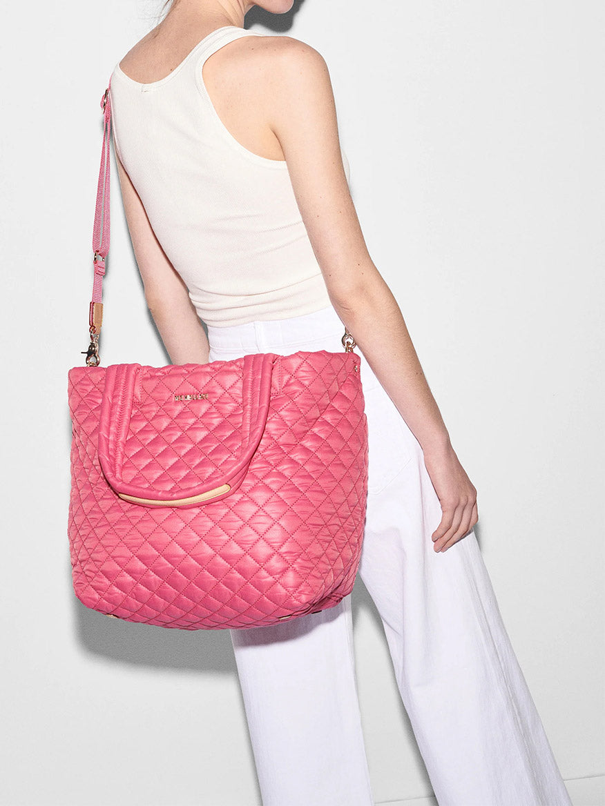 Woman holding a pink MZ Wallace Medium Metro Tote Deluxe in Zinnia Oxford, wearing a white tank top and white pants, against a grey background.