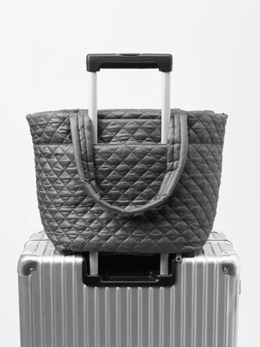 The MZ Wallace Medium Metro Tote Deluxe in Pewter Metallic Oxford, featuring a removable crossbody strap, is placed on top of a hard-shell suitcase with an extended handle.