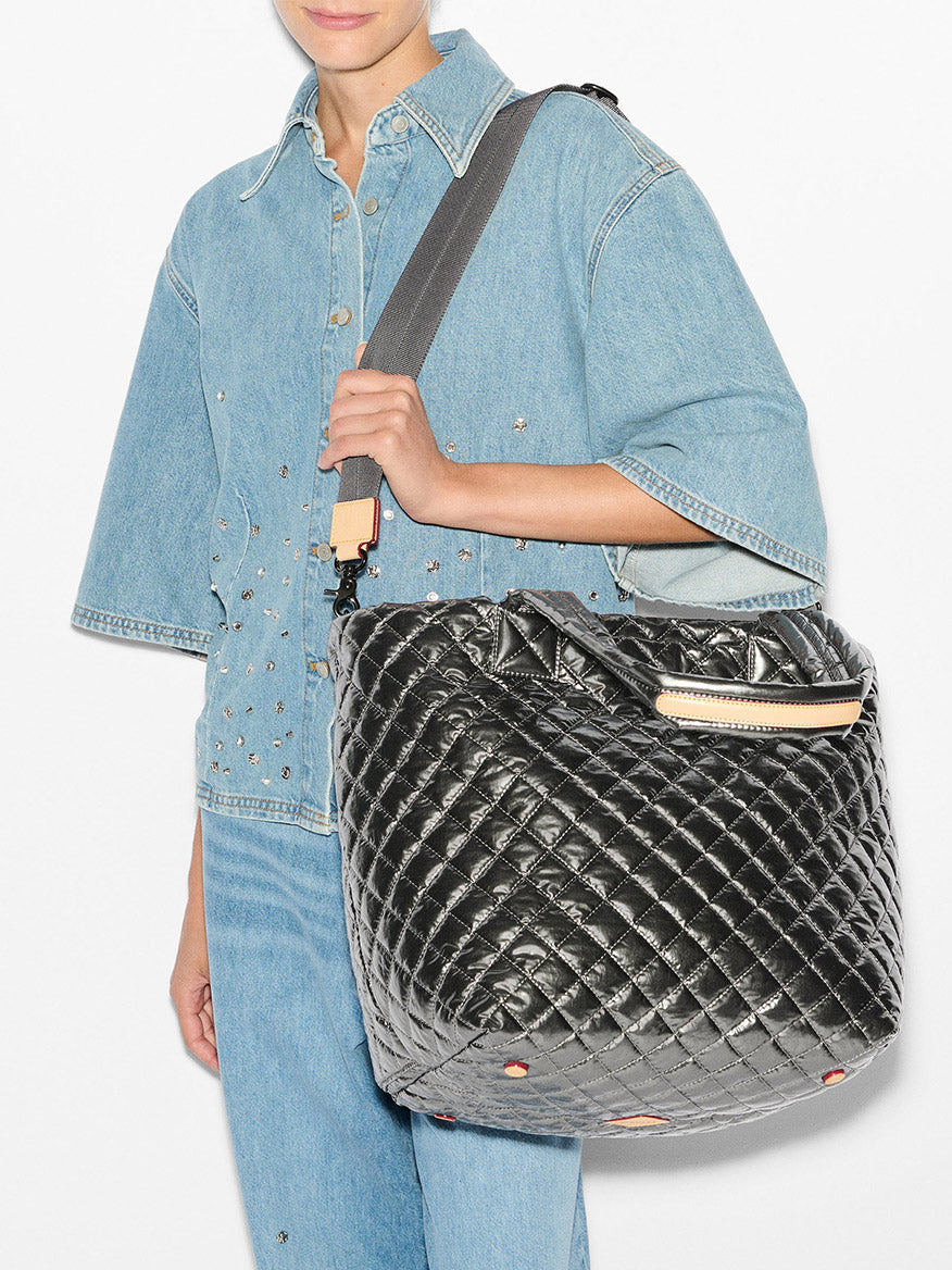 A person in a denim outfit holds the MZ Wallace Medium Metro Tote Deluxe in Pewter Metallic Oxford, featuring a large black quilted design with a removable crossbody strap.