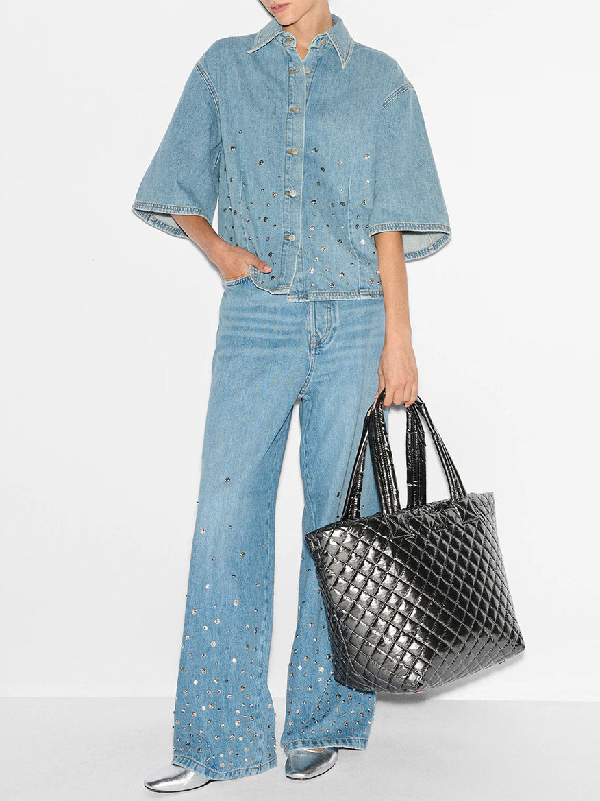 Person wearing a denim outfit with silver studs, holding an MZ Wallace Medium Metro Tote Deluxe in Pewter Metallic Oxford, featuring a removable crossbody strap and luggage sleeve, and wearing silver shoes.