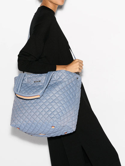 A person in a black outfit holds the MZ Wallace Medium Metro Tote Deluxe, a large quilted handbag in stone blue with Italian leather trim.