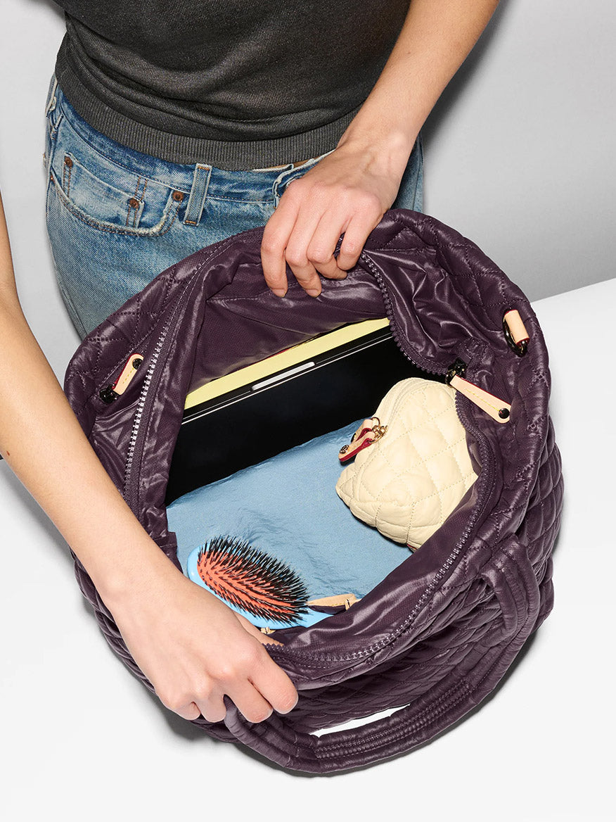 A person in jeans and a dark shirt opens an MZ Wallace Medium Metro Tote Deluxe in Raisin Oxford, revealing a laptop, blue cloth, beige quilted pouch, and hairbrush inside.