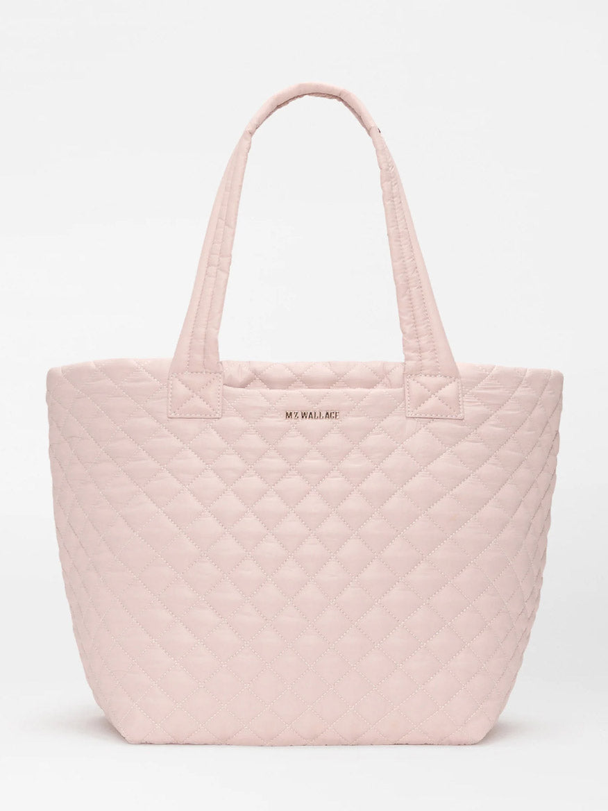 The MZ Wallace Medium Metro Tote Deluxe in City Pink Oxford is a light pink quilted bag with two handles, a luggage sleeve, "MZ WALLACE" at the top center, and is shown against a plain white background.