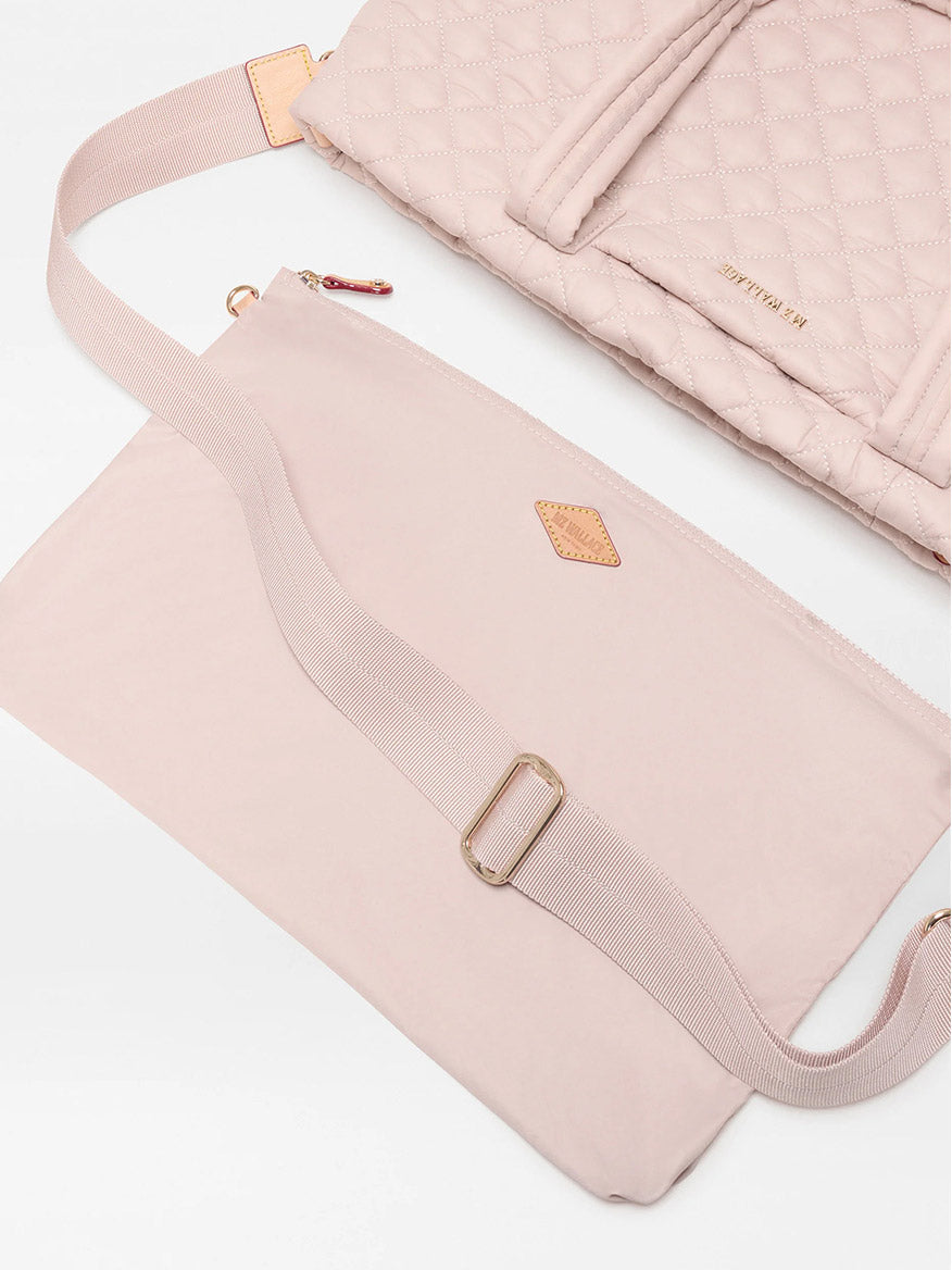 The MZ Wallace Medium Metro Tote Deluxe in City Pink Oxford, featuring a pale pink quilted texture and gold-tone hardware, sits above a matching flat pouch with an adjustable strap, both displayed on a white surface.