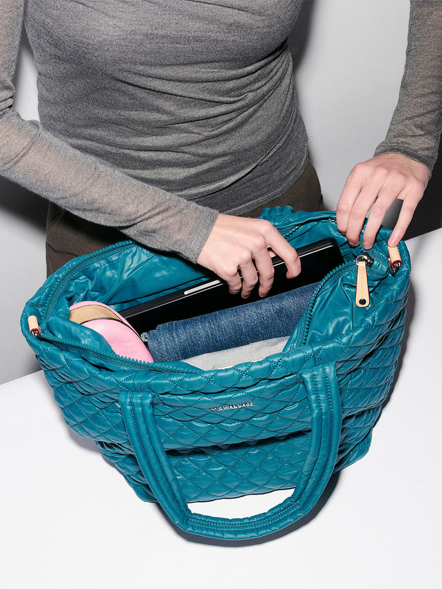 Someone places a laptop into the MZ Wallace Medium Metro Tote Deluxe in Pacific Oxford, which also holds a rolled-up pair of jeans and a pink item.