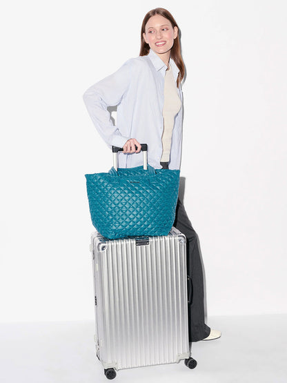 A woman stands beside a silver rolling suitcase with the MZ Wallace Medium Metro Tote Deluxe in Pacific Oxford on top, wearing a light shirt, grey pants, and cream shoes.