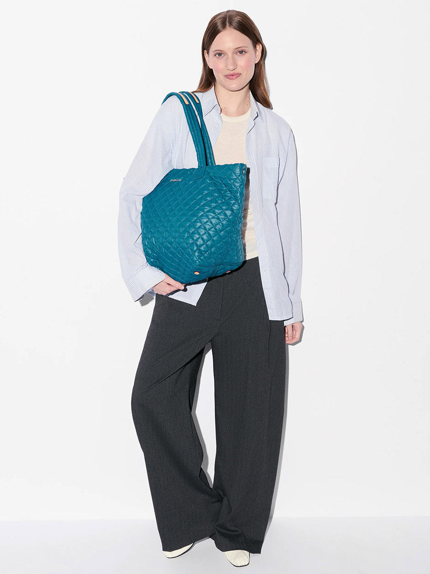 A woman stands against a white background, wearing a light blue shirt, cream top, dark wide-leg trousers, white shoes, and carrying the MZ Wallace Medium Metro Tote Deluxe in Pacific Oxford with quilted details.