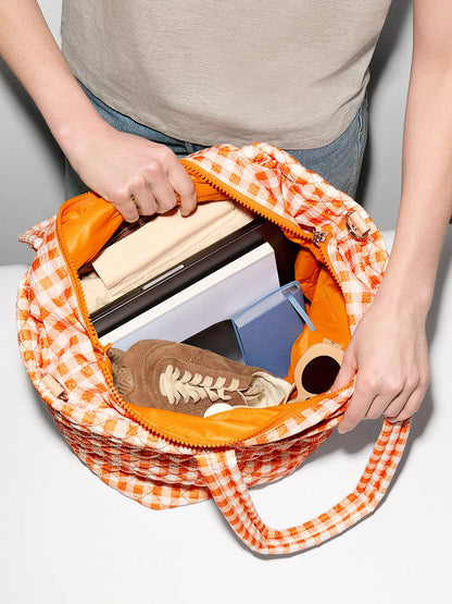 Someone opens an MZ Wallace Medium Metro Tote Deluxe in Sunset Orange Gingham Oxford, revealing a laptop, notebook, shoes, rolled-up shirt, and sunglasses inside.
