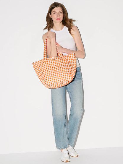 A woman in a white sleeveless top and blue jeans holds the MZ Wallace Medium Metro Tote Deluxe in Sunset Orange Gingham Oxford, standing against a plain white background.