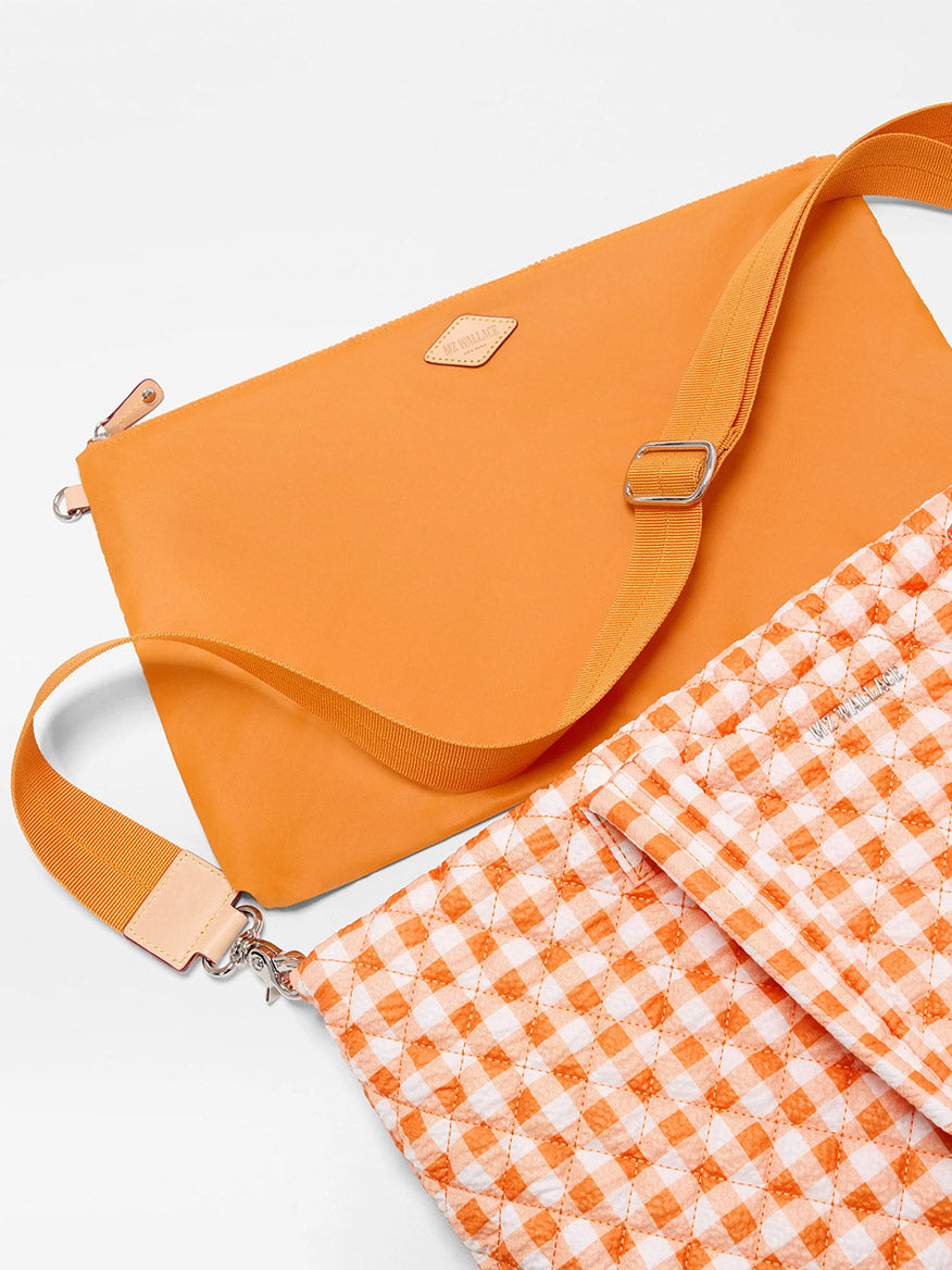The MZ Wallace Medium Metro Tote Deluxe in Sunset Orange Gingham Oxford, made from recycled nylon, features adjustable straps—one solid orange and one gingham—ideal for work and travel, displayed on a white surface.