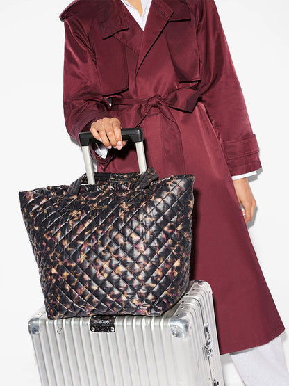 MZ Wallace Medium Metro Tote Deluxe in Tortoise Shell Oxford resting on a silver hard-shell suitcase.