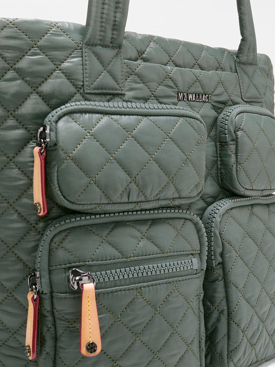 A close-up of the MZ Wallace Medium Metro Travel Tote in Juniper Oxford, crafted from recycled Oxford nylon, featuring a quilted green design, three zippered pockets, and an “MZ WALLACE” metal logo on the top pocket.