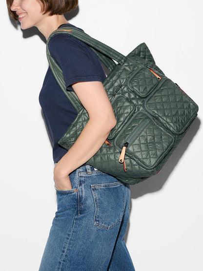 A person wearing a navy t-shirt and blue jeans carries the MZ Wallace Medium Metro Travel Tote in Juniper Oxford over one shoulder.