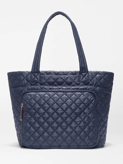 The MZ Wallace Medium Metro Travel Tote in Dawn Oxford is a navy blue quilted bag made from recycled nylon, featuring two handles and a front zippered pocket, shown against a plain white background.