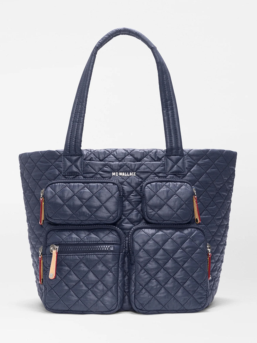 The MZ Wallace Medium Metro Travel Tote in Dawn Oxford is a navy blue quilted bag made from recycled nylon, featuring four front zippered pockets, long handles, and “MZ WALLACE” text on the front—ideal as a stylish laptop bag or everyday carryall.