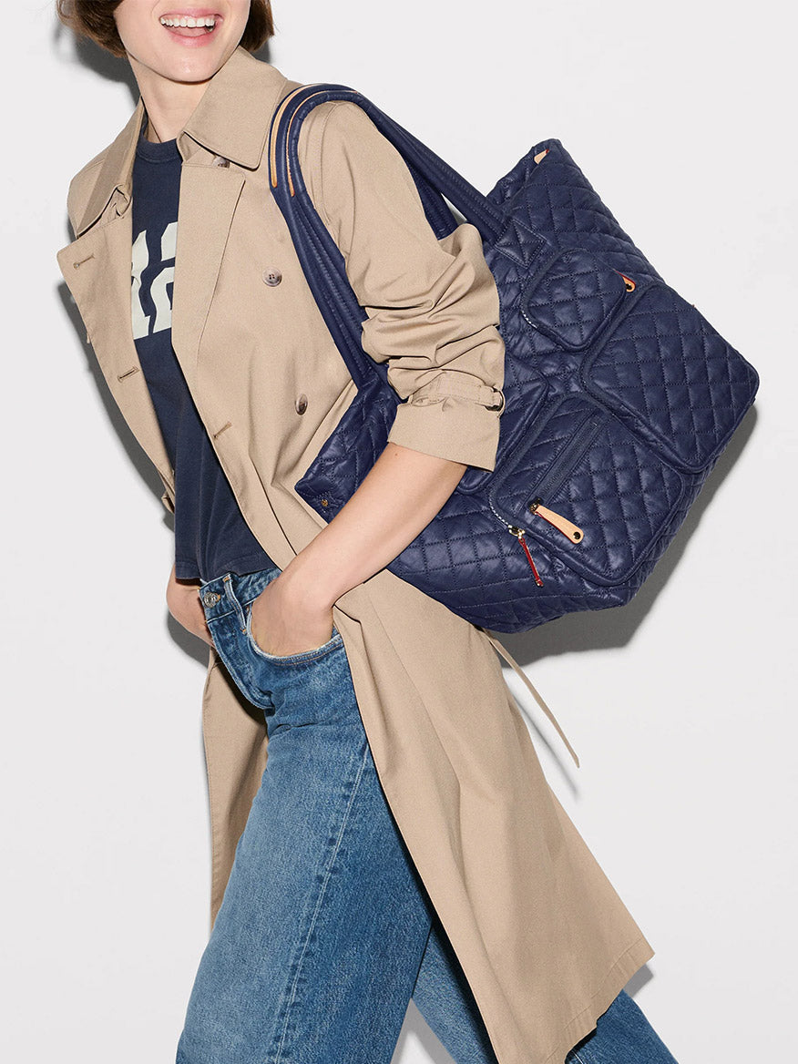 A person wearing a beige trench coat, blue jeans, and a navy t-shirt carries the MZ Wallace Medium Metro Travel Tote in Dawn Oxford—crafted from recycled nylon—on their shoulder against a plain background.