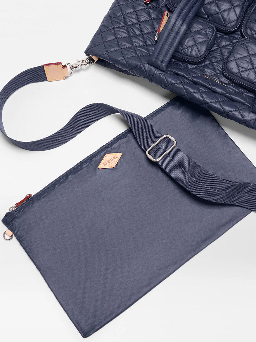 The MZ Wallace Medium Metro Travel Tote in Dawn Oxford, crafted from navy blue quilted fabric, is partially shown above a matching detachable recycled nylon pouch featuring an adjustable strap and a leather patch.