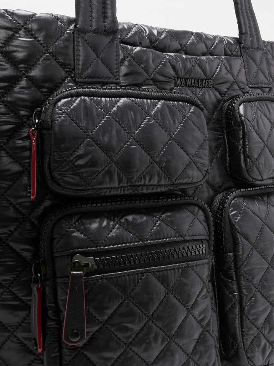 Close-up of the MZ Wallace Medium Metro Travel Tote in Black Oxford—a black quilted bag crafted from durable Black Oxford Nylon, featuring four front zippered pockets with red zipper pulls and a small MZ Wallace logo—making it an ideal choice for work and travel.