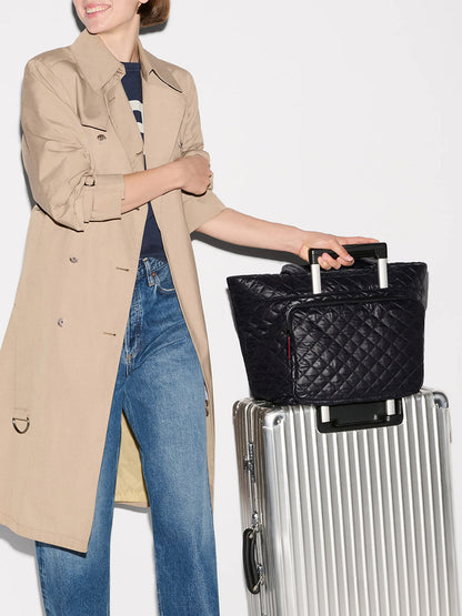 A person in a beige trench coat and jeans stands next to a silver suitcase with an MZ Wallace Medium Metro Travel Tote in Black Oxford on top, pulling the luggage handle.