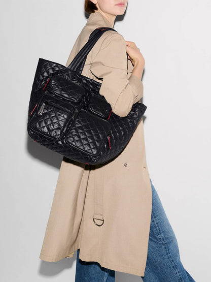 A person in a beige trench coat and blue jeans carries the MZ Wallace Medium Metro Travel Tote in Black Oxford, a spacious Work and Travel Bag with multiple zippered pockets, over their shoulder.