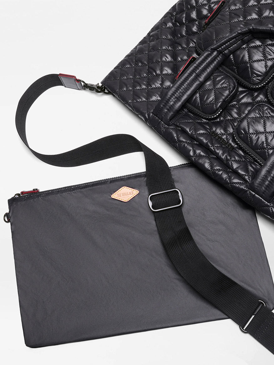 A MZ Wallace Medium Metro Travel Tote in Black Oxford with short handles is partially visible next to a flat black pouch featuring a shoulder strap and a small tan diamond-shaped logo tag.