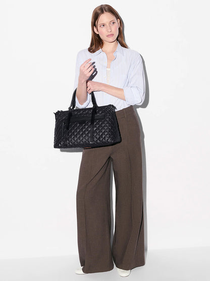 A woman stands against a white background wearing a light blue shirt, wide-leg brown trousers, white shoes, and holding the MZ Wallace Medium Park Satchel in Black Oxford—an ideal everyday bag.