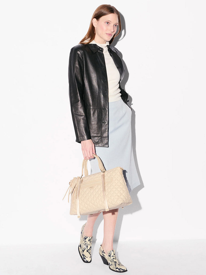 A woman stands against a white background wearing a black leather jacket, light skirt, white top, patterned ankle boots, and carrying the MZ Wallace Medium Park Satchel in Buff Oxford as her chic everyday bag.