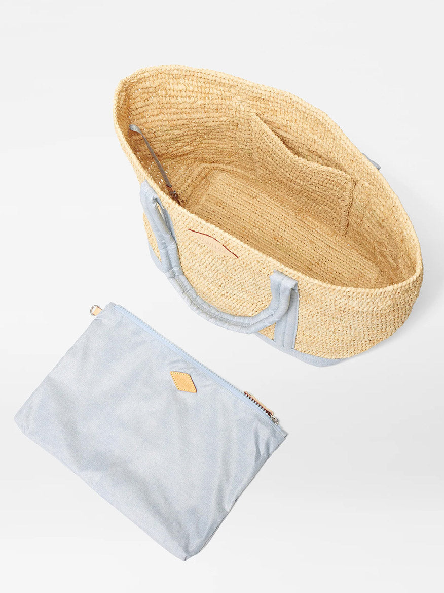 A woven straw MZ Wallace Medium Raffia Tote in Chambray/Raffia with light blue fabric trim and handles, accompanied by a matching light blue fabric pouch with a tan leather accent. Made from hand-crocheted Raffia, the tote features an interior pocket and is open at the top.