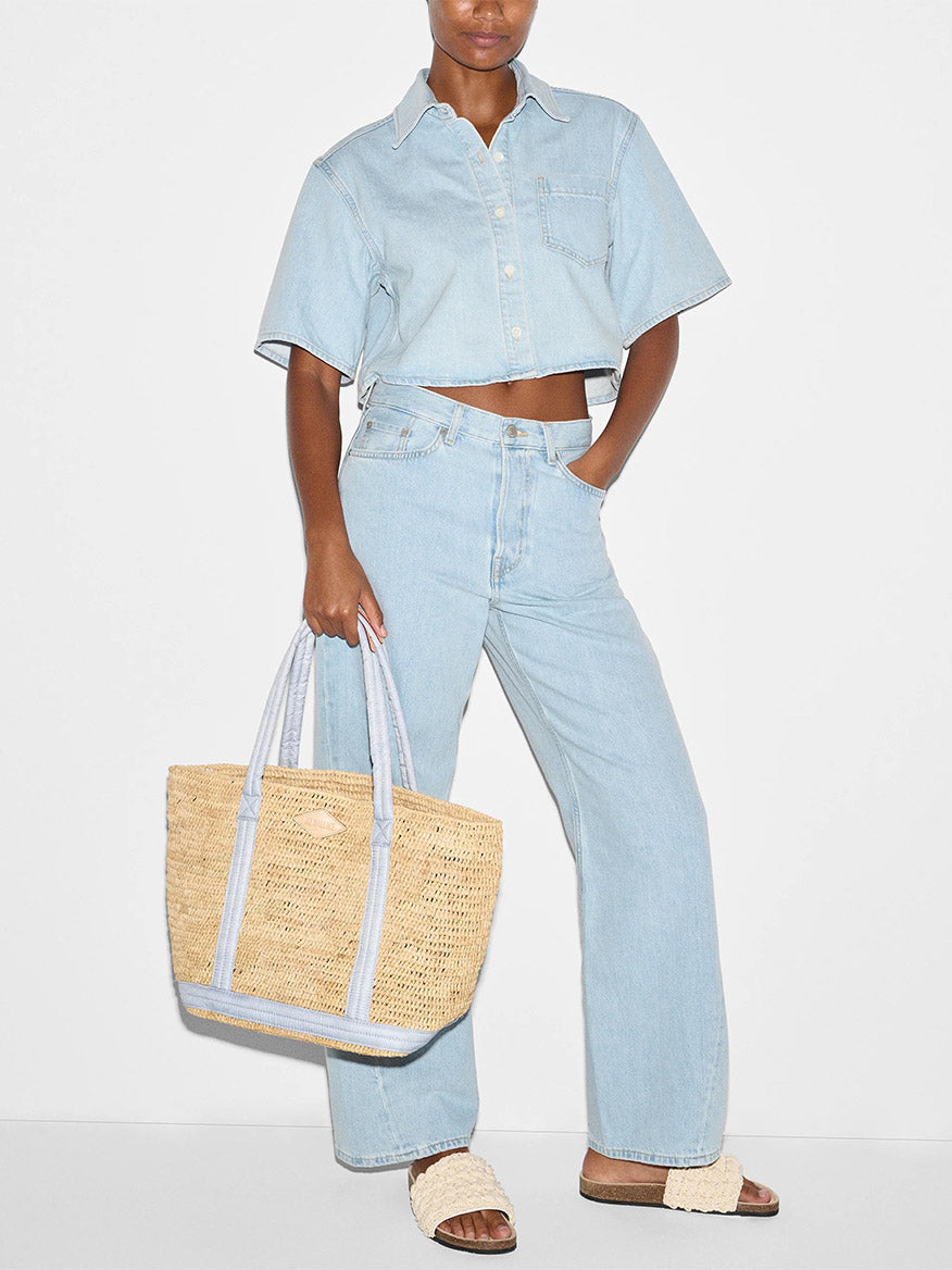 A person wearing a light blue denim crop shirt and matching jeans is holding an MZ Wallace Medium Raffia Tote in Chambray/Raffia and wearing slip-on sandals.