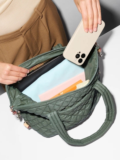 A person places a smartphone into an open MZ Wallace Medium Sutton Deluxe in Juniper Oxford, a quilted green handbag crafted from recycled nylon, which contains documents and a light blue item.
