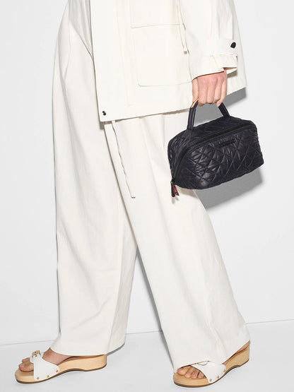 A person in loose white clothing and sandals holds the MZ Wallace Metro Accordion Cosmetic II in Black Oxford by its Italian leather trim handle.