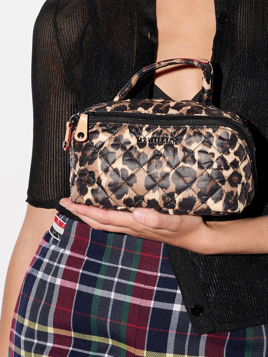 A person holds the MZ Wallace Metro Accordion Cosmetic II in Leopard Oxford, featuring a quilted leopard print and Italian leather trim, while wearing a black sheer top and plaid skirt.
