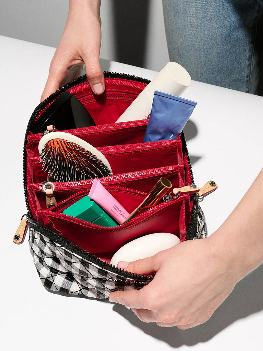A person holds the MZ Wallace Metro Accordion Cosmetic II in Black/White Gingham Oxford, using this stylish organizer with red lining to neatly store toiletries, makeup, and a hairbrush—perfect for travel essentials.