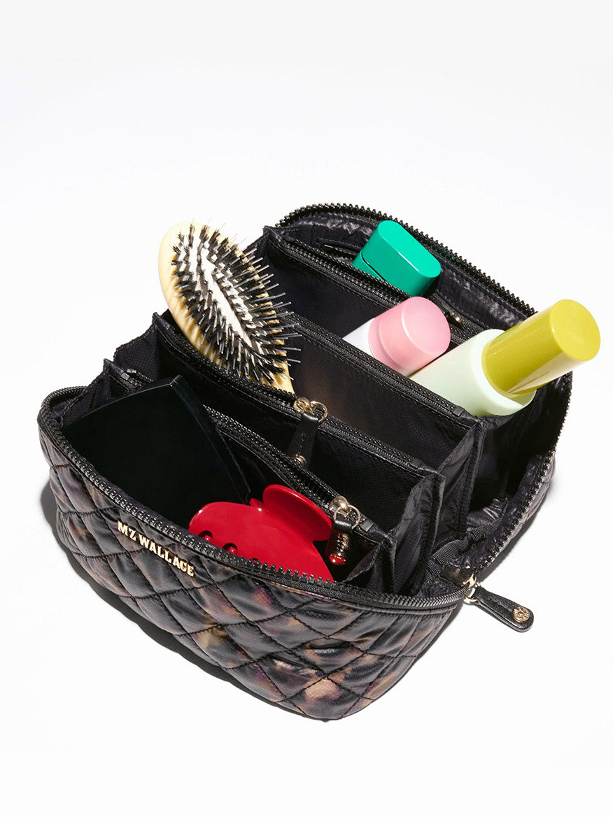 A black, quilted MZ Wallace Metro Accordion Cosmetic II in Tortoise Shell Oxford containing a hairbrush, several small bottles, and a red heart-shaped object.