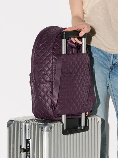 A person in a beige shirt and blue jeans rests the MZ Wallace Metro Backpack II in Raisin Oxford on the handle of a silver hard-shell suitcase.