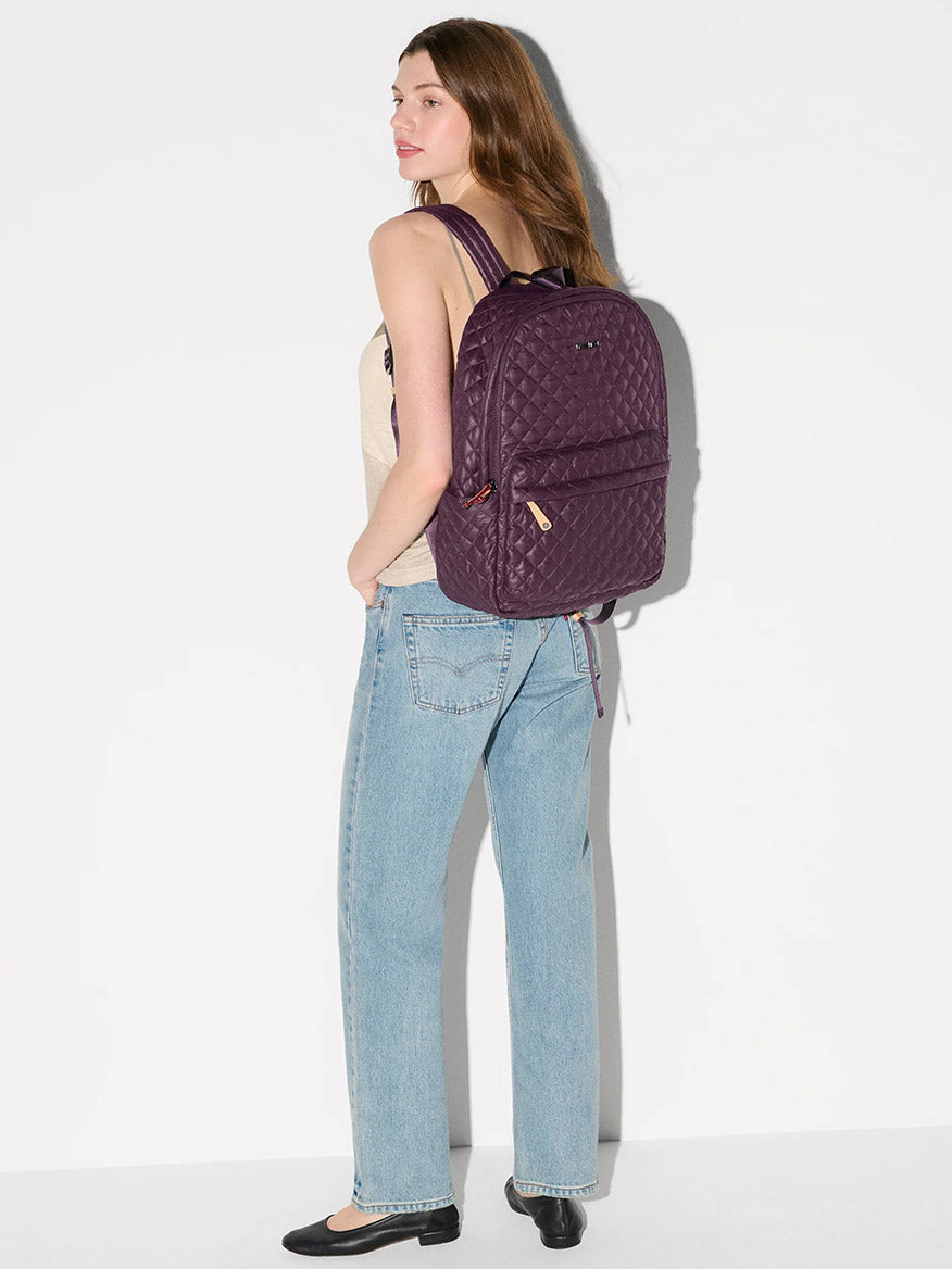 A woman stands sideways against a white background, wearing a beige tank top, light blue jeans, black flats, and carrying the MZ Wallace Metro Backpack II in Raisin Oxford.