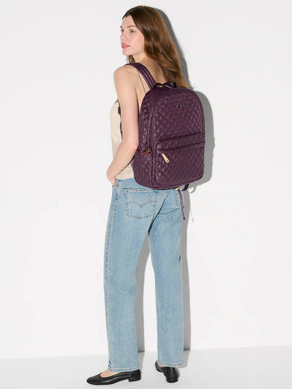A woman stands sideways against a white background, wearing a beige tank top, light blue jeans, black flats, and carrying the MZ Wallace Metro Backpack II in Raisin Oxford.