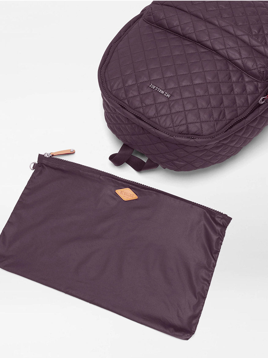 The MZ Wallace Metro Backpack II in Raisin Oxford, made from recycled Oxford nylon, and a matching zippered pouch are displayed on a white surface.