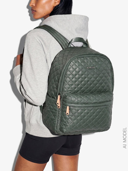 A person in a gray hoodie and black shorts wears the MZ Wallace Metro Backpack II in Juniper Oxford—a large, quilted green backpack with gold zippers crafted from recycled nylon—while standing against a plain light background.
