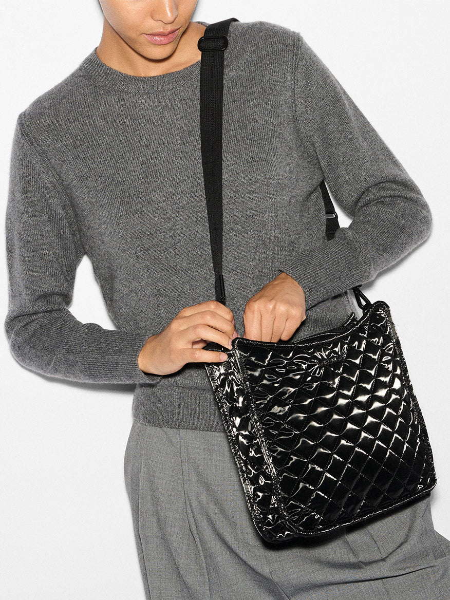 A person dressed in a gray sweater holds the MZ Wallace Metro Box Crossbody in Black Liquid Oxford, its quilted texture adding an elegant touch.